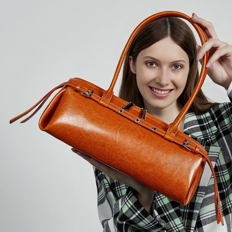A woman holding a structured cylindrical orange-brown leather handbag with dual top handles and a long shoulder strap, posed against a light gray studio background.