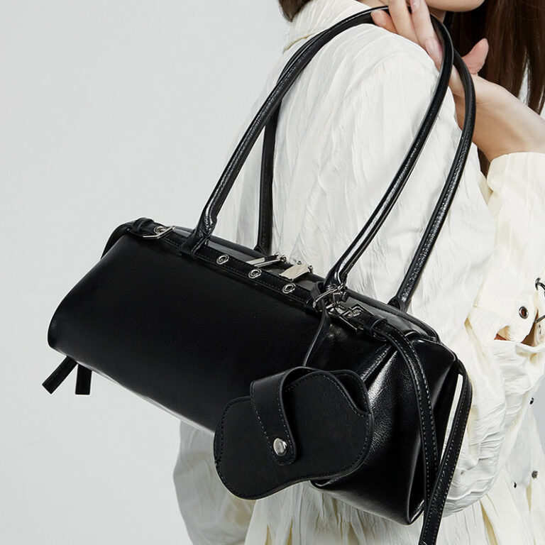 A black leather shoulder bag with dual top handles and a small matching pouch, shown being worn over the shoulder of a person in a white blouse against a light gray background.