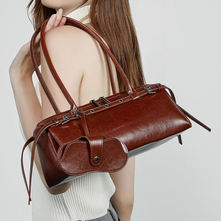 A woman wearing a white ribbed top carries a brown faux-leather shoulder bag with a structured rectangular shape, double top handles, and a front flap pocket with a snap closure, against a plain light gray background.